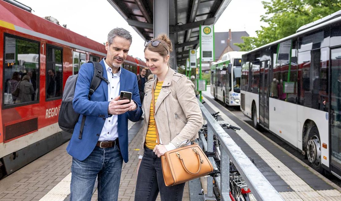 Eine Frau und ein Mann stehen am Bahnsteig und schauen auf ein Smartphone. Links hält eine Bahn, rechts zwei Busse.