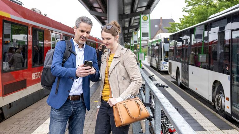 Eine Frau und ein Mann stehen am Bahnsteig und schauen auf ein Smartphone. Links hält eine Bahn, rechts zwei Busse.